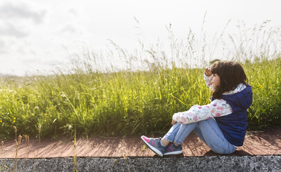Kleines Mädchen sitzt auf dem Bürgersteig vor einer Wiese und schaut in die Ferne - MGOF001934