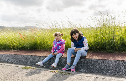 Two laughing little sisters sitting at roadside stock photo