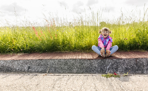 Lachendes kleines Mädchen auf dem Bürgersteig vor einer Wiese sitzend, lizenzfreies Stockfoto