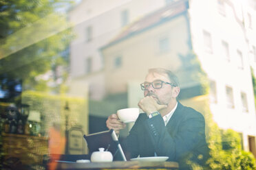 Pensive businessman with cup of coffee in a cafe behind windowpane - MAEF011819