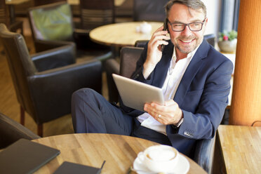Portrait of smiling businessman with digital tablet sitting in a cafe telephoning with smartphone - MAEF011820