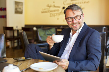 Porträt eines lächelnden Geschäftsmannes mit digitalem Tablet, der in einem Café sitzt und Kaffee trinkt - MAEF011822