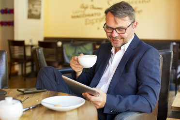 Portrait of smiling businessman using digital tablet while sitting in a cafe drinking coffee - MAEF011823