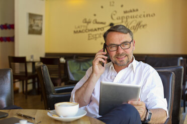 Porträt eines lächelnden Geschäftsmannes mit digitalem Tablet, der in einem Café sitzt und mit einem Smartphone telefoniert - MAEF011828