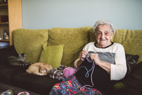 Portrait of crocheting senior woman sitting on couch besides her sleeping cats - RAEF001202