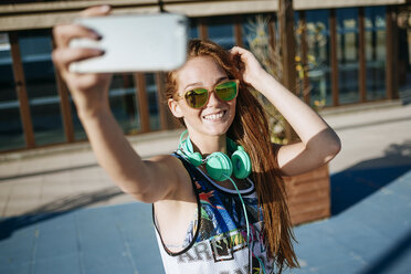 Lächelnde junge Frau mit verspiegelter Sonnenbrille macht Selfie mit Smartphone - KIJF000462