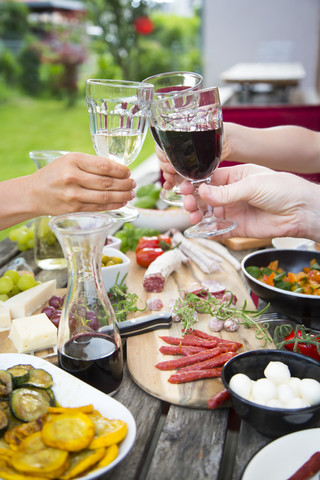 Menschen stoßen mit Weingläsern an und genießen eine Vielzahl von mediterranen Antipasti im Garten, lizenzfreies Stockfoto