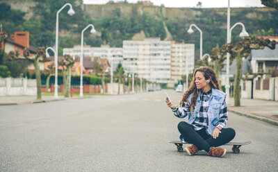 Junge Frau hört Musik und sitzt auf einem Skateboard - DAPF000131