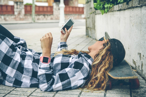 Junge Frau liegt auf einem Skateboard und schaut auf ihr Handy, lizenzfreies Stockfoto