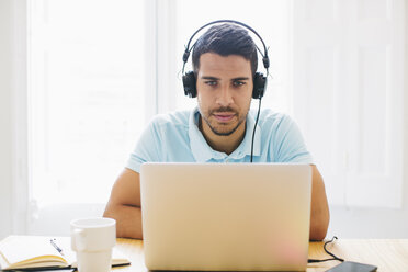 Junger Mann im Büro, der Kopfhörer trägt und einen Laptop benutzt - EBSF001530