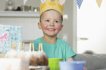 Glücklicher Junge mit Papierkrone, der am Geburtstagstisch sitzt - NHF001500