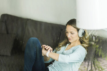 Woman at home on couch wearing headphones looking at smartwatch - SBOF000099