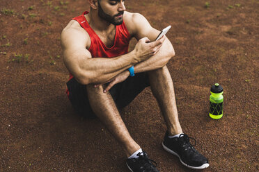 Athlete outdoors sitting on ground looking at cell phone - UUF007725