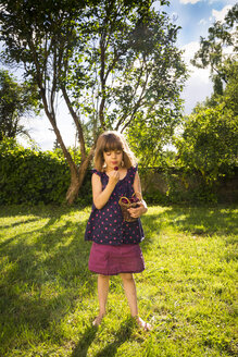 Kleines Mädchen steht auf einer Wiese im Garten und betrachtet eine Kirsche - LVF004963