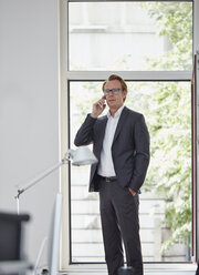 Businessman in his office standing in front of open window telephoning with smartphone - RHF001599