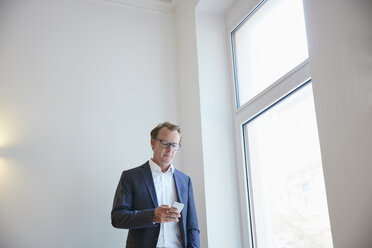 Businessman standing beside window looking at his smartphone - RHF001622