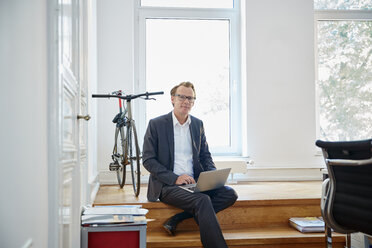 Businessman with laptop sitting on steps in an office - RHF001631