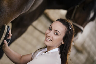 Porträt einer lächelnden jungen Frau, die ein braunes Pferd striegelt - MIDF000726