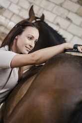 Junge Frau striegelt den Rücken eines braunen Pferdes - MIDF000727