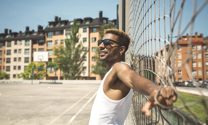 Junger Mann mit Sonnenbrille lehnt am Netz des Basketballplatzes - DAPF000183