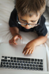 Kleiner Junge mit Brille liegt auf dem Bett und benutzt einen Laptop - VABF000577