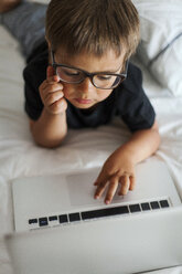 Little boy wearing glasses lying on bed using laptop - VABF000578