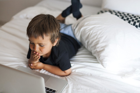 Lächelnder kleiner Junge auf dem Bett liegend mit Laptop, lizenzfreies Stockfoto