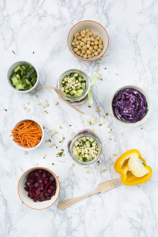 Zwei Gläser Regenbogensalat mit Kichererbsen, Sprossen und verschiedenen Gemüsesorten und Schalen mit Zutaten, lizenzfreies Stockfoto