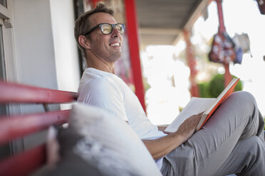 Lächelnder Mann sitzt auf Veranda und liest ein Buch - ZEF008799