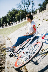 Young woman with bicycle sitting on tilted wall - GIOF001214