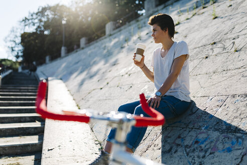 Junge Frau mit Fahrrad und Kaffee zum Mitnehmen sitzt auf einer schiefen Mauer - GIOF001224