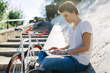 Junge Frau mit Fahrrad, die an einer schrägen Wand sitzt und einen Laptop benutzt - GIOF001227