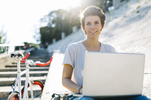 Junge Frau mit Fahrrad, die im Freien sitzt und einen Laptop benutzt, lizenzfreies Stockfoto