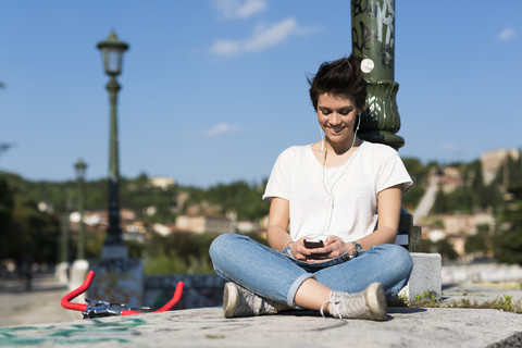 Lächelnde junge Frau sitzt an der Wand und hört Musik, lizenzfreies Stockfoto