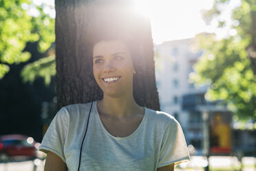 Lächelnde junge Frau am Baumstamm mit Kopfhörern - GIOF001242