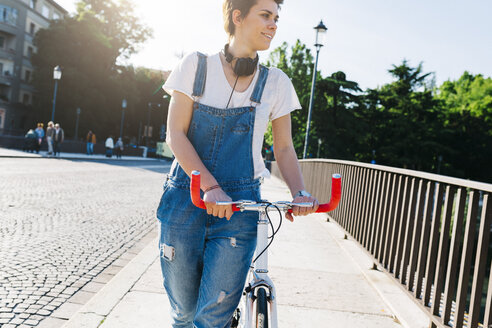 Lächelnde junge Frau, die ein Fahrrad auf einer Brücke schiebt - GIOF001244