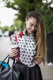 Portrait of smiling woman with coffee to go looking at her smartphone - MAUF000662