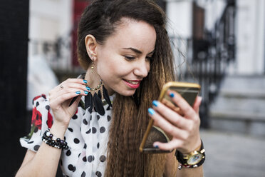 UK, London, young woman watching her earring with the aid of her smartphone - MAUF000670