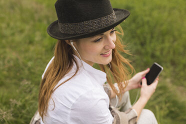 Junge Frau auf einer Wiese, die mit Kopfhörern und Smartphone Musik hört - BOYF000443