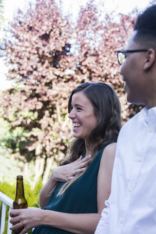 Lachende Frau, die bei einem sommerlichen Abendessen in einem Garten eine Flasche Bier trinkt, lizenzfreies Stockfoto