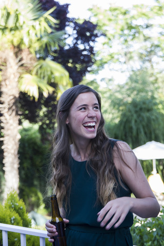 Lachende Frau mit einer Bierflasche in der Hand bei einem sommerlichen Abendessen in einem Garten, lizenzfreies Stockfoto