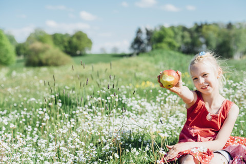 Lächelndes Mädchen sitzt auf einer Wiese und hält einen Apfel, lizenzfreies Stockfoto