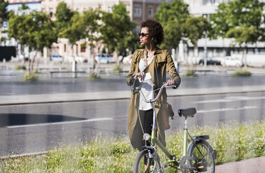 Junge Frau mit Smartphone und Kopfhörern, die ein Fahrrad schiebt - UUF007734