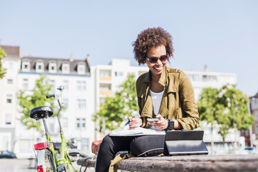 Lächelnde junge Frau, die auf einer Bank sitzt und ein Smartphone und ein digitales Tablet benutzt - UUF007736