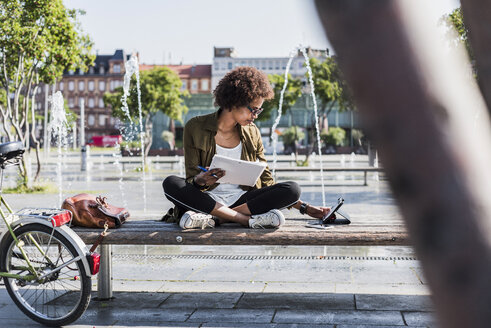 Young woman sitting on a bench with notebbok looking at digital tablet - UUF007739
