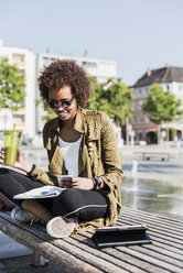 Junge Frau sitzt mit Getränk und Smartphone auf einer Bank und liest Notizen - UUF007741