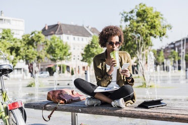 Junge Frau sitzt auf einer Bank und trinkt ein Getränk, während sie auf ihr Smartphone schaut - UUF007742