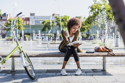 Young woman sitting on a bench with beverage reading notes - UUF007748