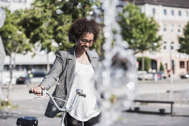 Porträt einer lächelnden Frau mit Fahrrad, die mit Kopfhörern Musik hört - UUF007751