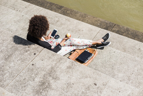 Junge Frau sitzt auf einer Treppe und isst einen Apfel, während sie auf ihr Smartphone schaut - UUF007764